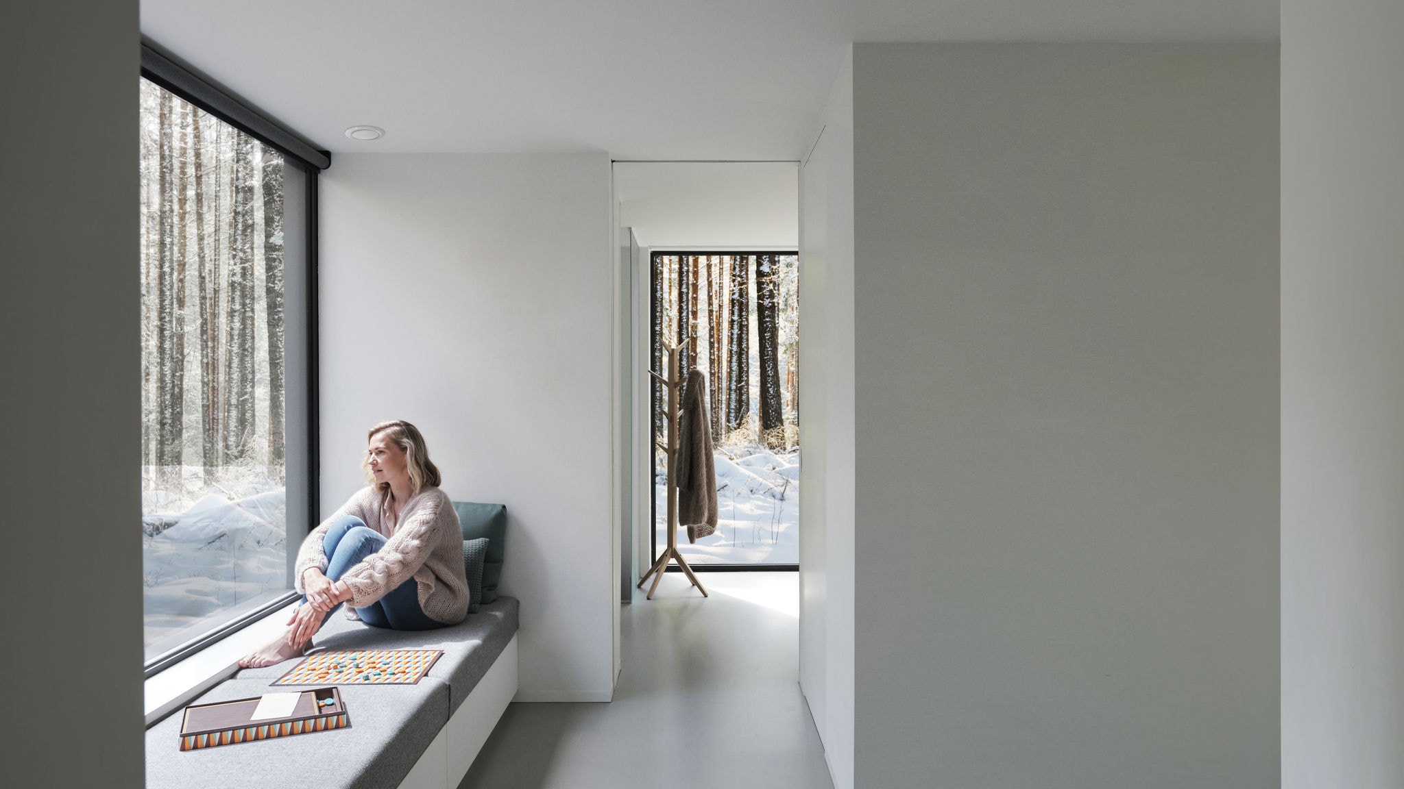 Weitwinkelaufnahme einer Frau, die barfuß vor einem Fenster sitzt und auf die Bäume und den Schnee hinausschaut.