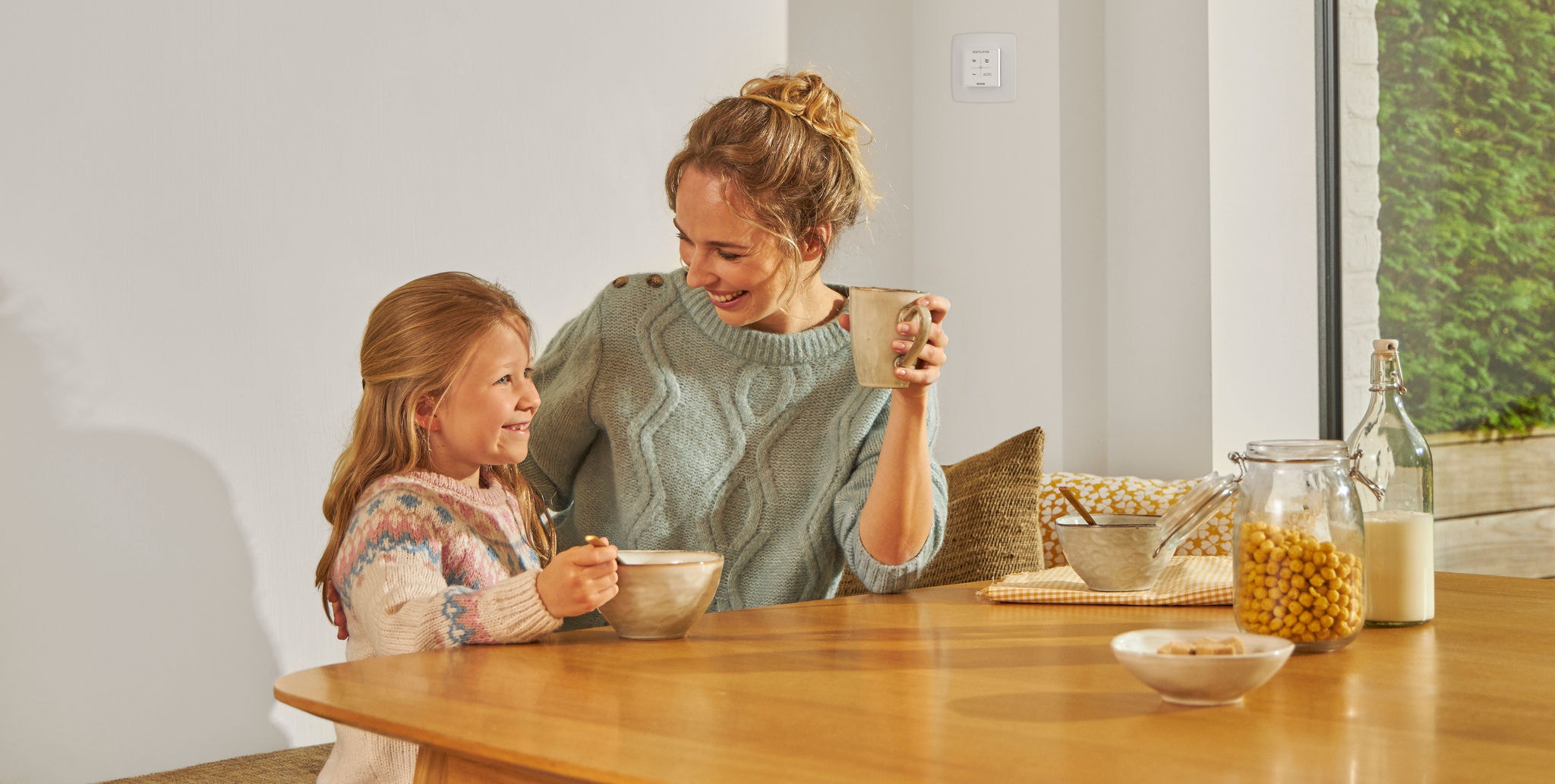 Ein junges Mädchen sitzt aus einer Schüssel essend an einem Tisch neben einer Frau mit einer Tasse in der Hand; beide lächeln, sind glücklich und entspannt und genießen die gesunde Luft.