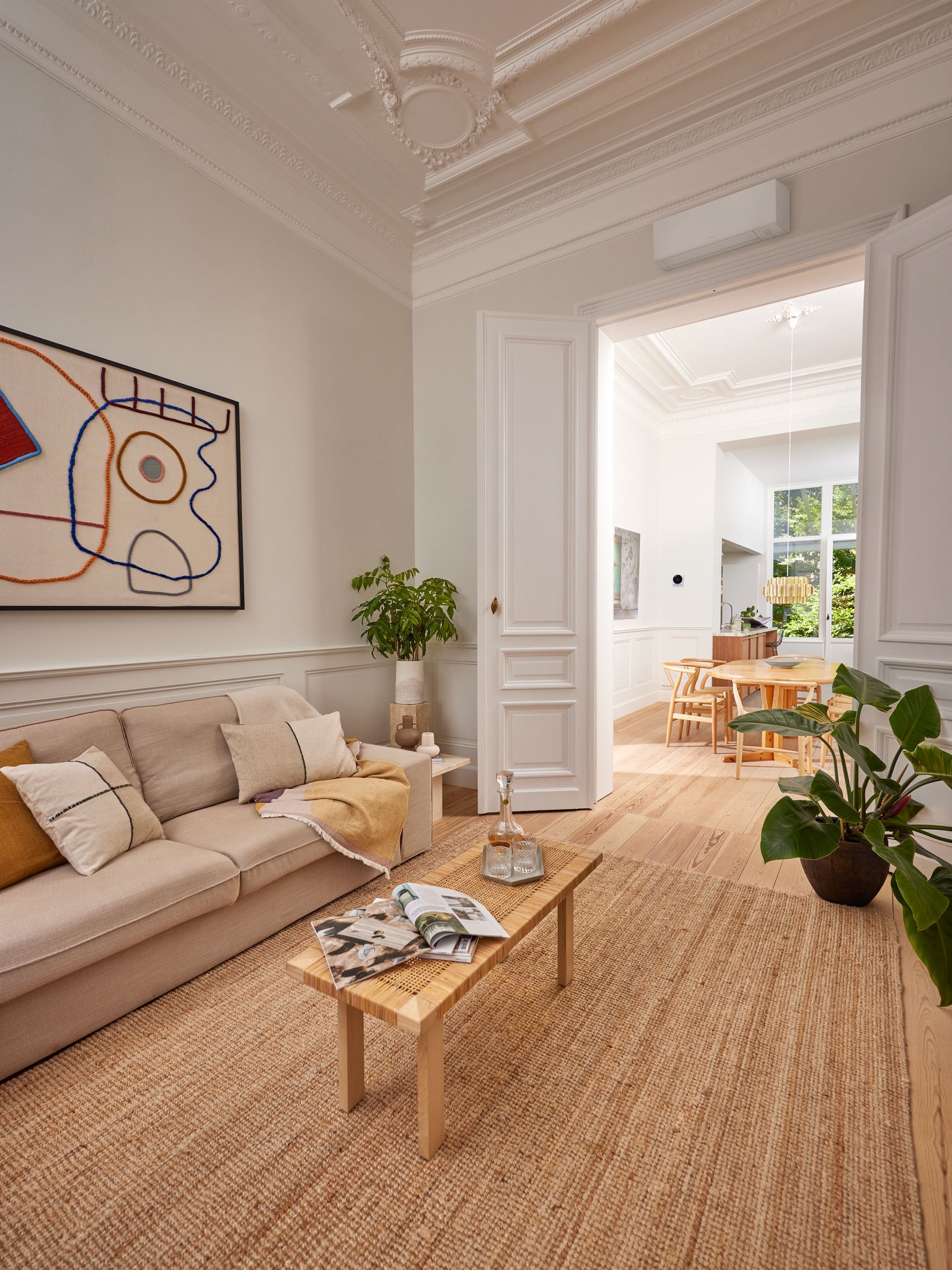 Weißes Wärmepumpen-Wandgerät über einer Tür, die von einem behaglichen Wohnzimmer mit cremefarbenem Sofa in ein durch ein großes Fenster sonnenüberflutetes Esszimmer führt