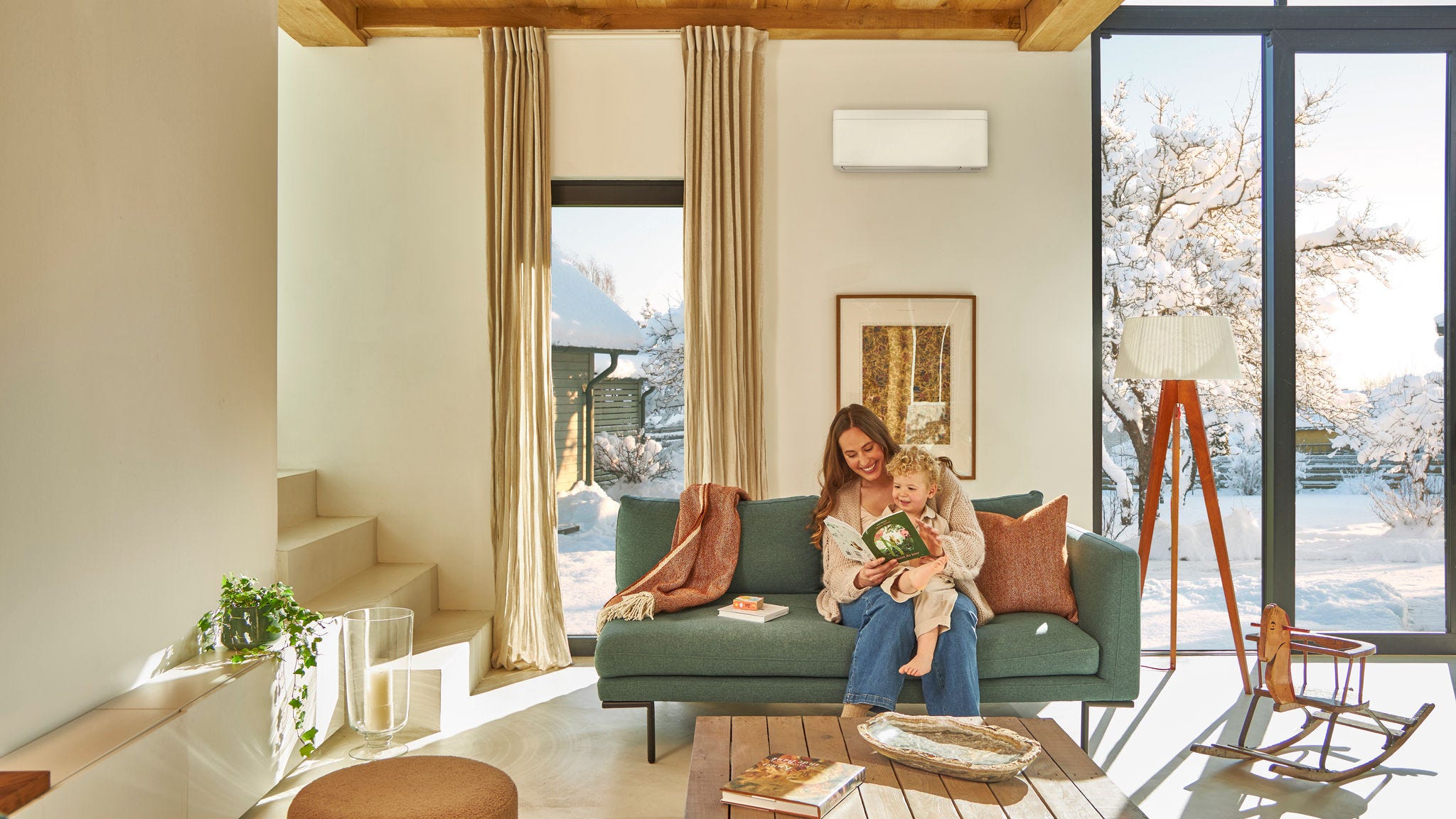 Unter einem Wandgerät auf der Couch sitzende Mutter liest einem auf ihrem Schoß sitzenden Kleinkind ein Kinderbuch vor und genießt die Wärme, während es draußen schneit